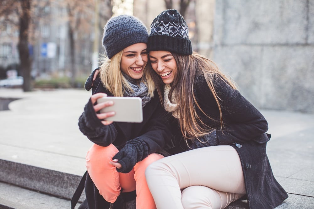 Two young women on bench look at best financial apps for teens and young adults on an iPhone